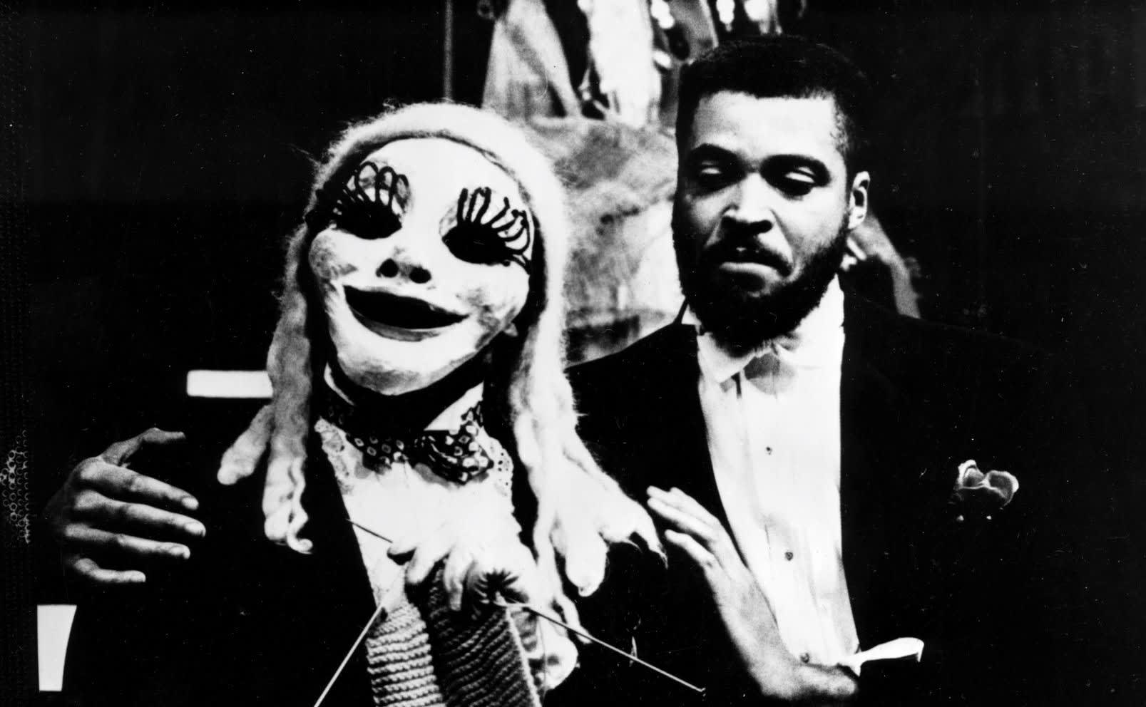 James Earl Jones has his hands on the shoulders of a person with a mask and wig who is knitting
